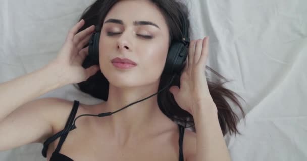 Mujer acostada en la cama, sostiene los auriculares, se balancea hacia la música, mira con sonrisa a la cámara — Vídeo de stock