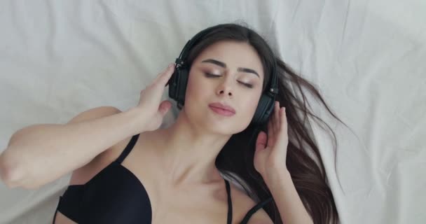 Girl listening to music on bed, looks into camera and twists headphones wire — Stock Video