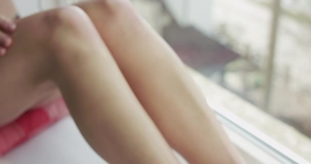 Tunnistamaton alaston nainen istuu ikkunalaudalla ja hyväilee jalkojaan — kuvapankkivideo