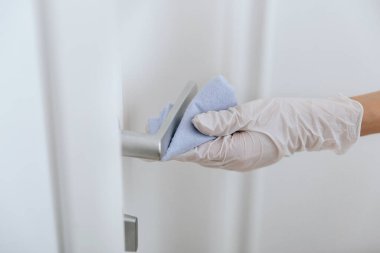 Beyaz eldivenli, mavi mendilli kapı kolu. Ev kapısı bağlantısını temizlemek için havlu kullanan kadın eli. Hastane ve halka açık alanlarda korona virüsüne karşı yüzeylerin sterilize edilmesi.