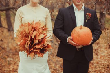 Sonbahar düğünü. Damat portakal kabağı tutuyor, gelin de bir buket sonbahar yaprağı tutuyor. Balkabaklı yaprak. Yüksek kalite fotoğraf. Şükran Günü veya Cadılar Bayramı konsepti afişi veya kartpostal.