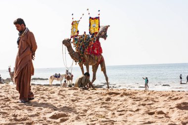 Bazı yerliler develerini ve atlarını bekleyen müşterileri bekliyor ve diğerleri de Pakistan 'daki Hawks Bay Karachi, Tushan Sahili' nde plajın tadını çıkarıyorlar.