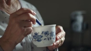 Yaşlı bir kadının bir fincan çayı kaşıkla karıştırıp içtiği bir fotoğraf.