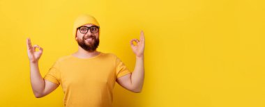Gözlüklü, gülümseyen sakallı adam sarı arka planda jest yapıyor, panoramik düzende.