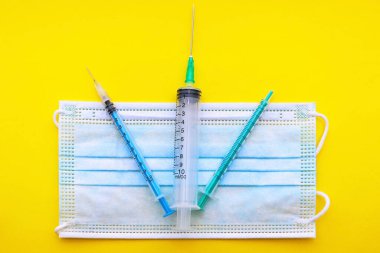 Tıbbi şırıngalar sarı arka planda, maskenin üzerinde tepeden tırnağa uzanır. konsept. Covid 19. Aşı olmadan.