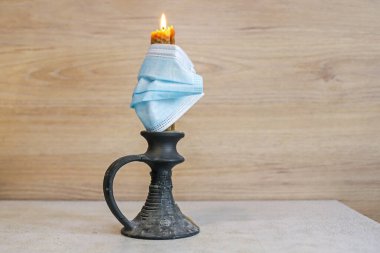 Üzerinde tıbbi maske olan bir şamdanın içinde yanan bir mum, ahşap bir duvarın arka planında masanın üzerinde duruyor.