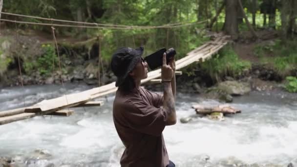 Boldog fickó, analóg kamerával a folyóparton áll a hegyekben. Férfi fotós, professzionális kamerával fényképez. Fiatal férfi látszó képek 8 mm-es kamera szabadban — Stock videók