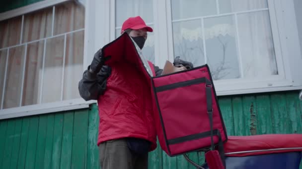 Un mensajero con uniforme rojo, máscara y guantes entrega un paquete. Trabajar en una pandemia — Vídeos de Stock