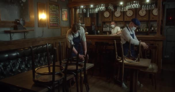 Barman a personál v obličejových maskách připravují restauraci na znovuotevření po karanténě. Mladí číšníci v zástěrách ochranné masky a rukavice uspořádání nábytku v kavárně před otevřením. The concept of — Stock video