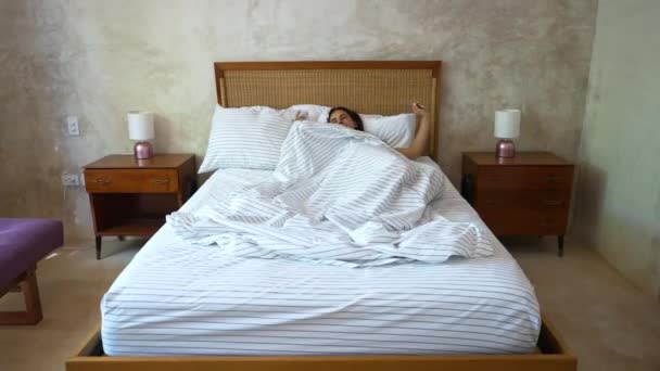 Šťastný ospalý mladý dospělý žena probuzení ležící v pohodlné bílé posteli. Pozitivní hezká dívka protahování těší brzy ráno v útulné ložnici. Čerstvá líná dáma se probouzí po zdravém spánku doma — Stock video