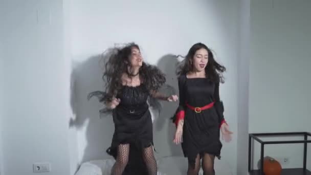 Dos chicas colombianas felices con disfraces negros de Halloween saltando en la cama en el apartamento. - Tiro medio — Vídeo de stock
