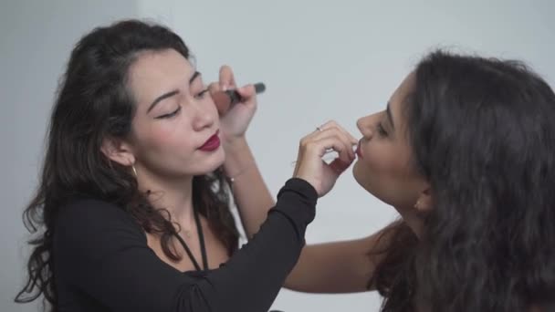 Vista lateral de duas garotas colombianas bonitas aplicando maquiagem umas com as outras enquanto se sentam na cama isoladas contra fundo de parede branca, Valência, Espanha. - tiro de close-up — Vídeo de Stock