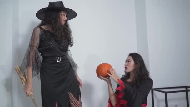 Две колумбийские девушки в костюмах ведьм, стоящие на кровати с одной, держащей метлу, и другая, держащая тыкву внутри квартиры в Валенсии, Испания. - Средний крупный план — стоковое видео