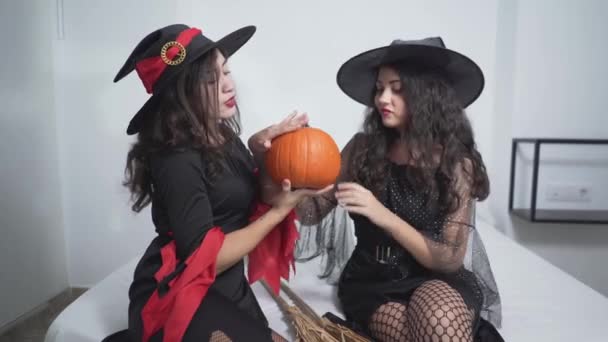 Twee mooie vrouwen dragen zwarte heksenjurk en hoed, die een pompoen vasthouden met hun beide handen terwijl ze op het bed van een appartement zitten in Valencia, Spanje. - Medium close-up schot. — Stockvideo