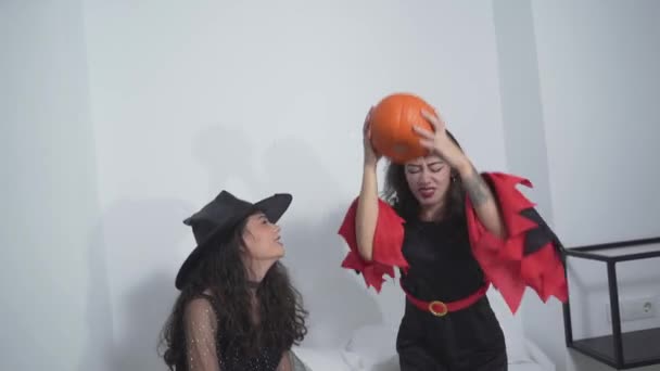 Dos mujeres alegres con disfraces negros de Halloween divirtiéndose con una calabaza en la cama. tiro completo — Vídeo de stock