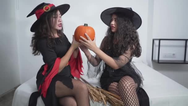 Két fiatal kolumbiai hölgy boszorkányjelmezben, akik egy narancssárga tökkel viccelődnek a szobában. közepes lövés — Stock videók