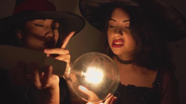 Dwie Kolumbijki w halloweenowym kostiumie noszące kapelusz czarownicy poruszające rękami wokół przezroczystej żarówki w ciemnym pokoju mieszkania w Walencji w Hiszpanii. - Strzał z bliska — Wideo stockowe