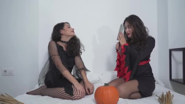 Twee mooie Colombiaanse meisjes gekleed in Halloween kostuum zitten op bed met pompoen, een meisje vasthouden spelen met een mes. Halloween Concept. - Middelmatig schot. — Stockvideo