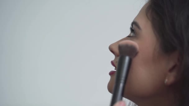 Hermosa dama colombiana mirando hacia arriba y sonriendo mientras el artista del maquillaje aplica rubor en su mejilla. primer plano, vista lateral — Vídeos de Stock