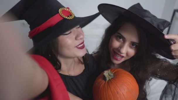 Retrato de dos encantadoras chicas colombianas vestidas con disfraces de bruja con una calabaza tomando foto dentro del apartamento en Valencia, España. - Primer plano, tiro de ángulo alto — Vídeo de stock