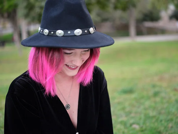 Mujer joven con el pelo rosado sonriendo y mirando hacia abajo, usando un sombrero y una blusa negra — Foto de Stock
