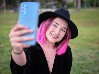 Siyah şapkalı ve pembe saçlı bir kadın parkta selfie çekiyor.