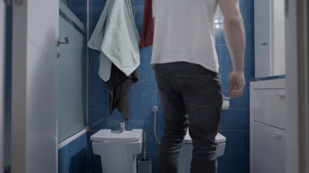 Ung kaukasier man kommer i badrummet och få ett nyhetsbrev för att sitta i toaletten - Frontal shot — Stockvideo
