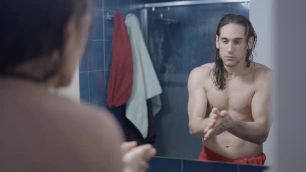 Jonge metroseksuele man met behulp van een serum voor zijn haar in de voorkant van de badkamer spiegel na een douche - Midden schot — Stockvideo