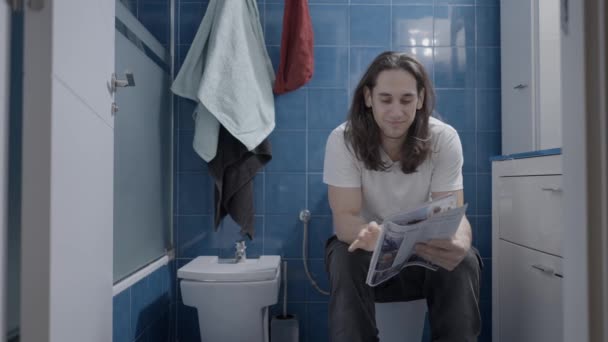 Young caucasian man reading a newsletter in the bathroom while shit - Frontal shot — Stock video