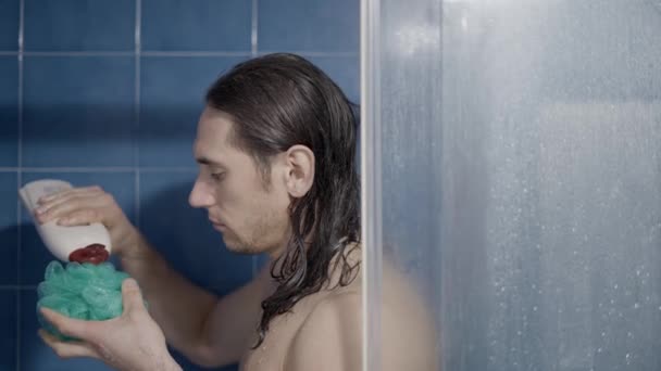 Fiatal, hosszú hajú férfi, aki fürdõszappant tölt egy fürdõszivaccsal a zuhany alatt - közelkép — Stock videók