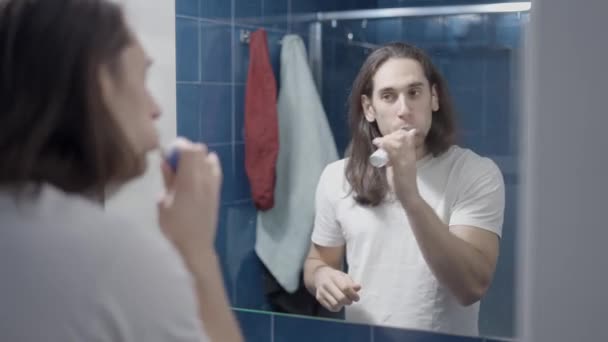 Jonge blanke man poetsen zijn tanden in de voorkant van een badkamer spiegel - Midden schot — Stockvideo