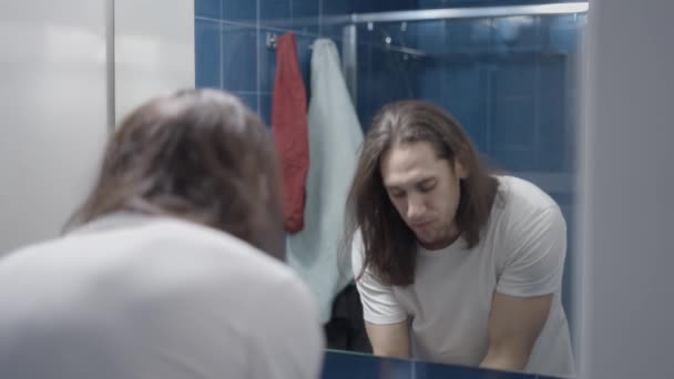 Длинноволосый мужчина, глядя на зубы сразу после чистки перед зеркалом в ванной - Медленное движение — стоковое видео