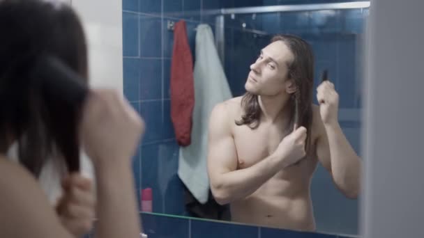 Jeune homme nu et aux cheveux longs se brossant les cheveux devant le miroir de la salle de bain - Plan du milieu — Video