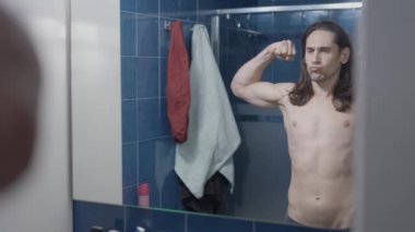 Genç çıplak adam banyo aynasının önünde kaslarına bakıyor.
