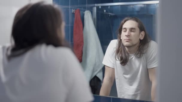 Молодой человек смотрит на себя в зеркало и думает о своем дне - Средний выстрел — стоковое видео