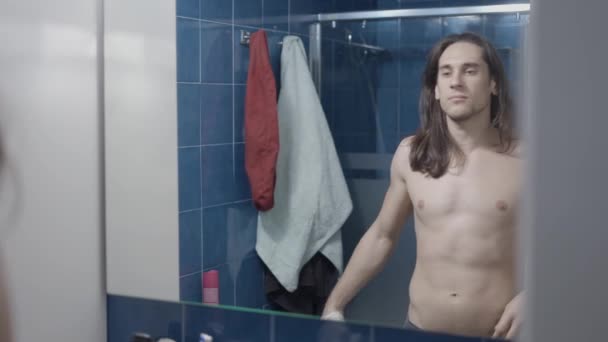 Jongeman neemt zijn shirt uit in de voorkant van zijn badkamer spiegel - Midden schot — Stockvideo