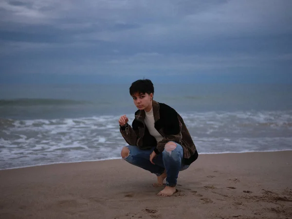 Trans woman squatting with the beach behind her, posing in warm clothes — 图库照片