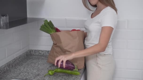 Mujer joven sacando verduras de la bolsa de papel de supermercado en la cocina. - tiro medio — Vídeos de Stock