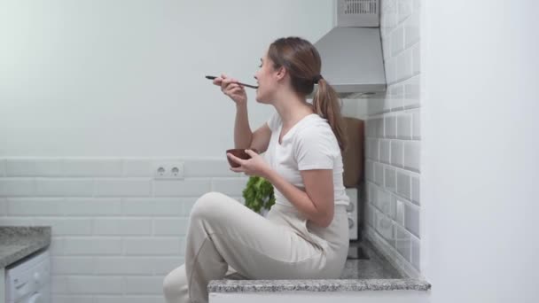 Cheerful Girl Eating While Sitting On Top Of Kitchen Countertop. Static, Sideview — Vídeo de Stock
