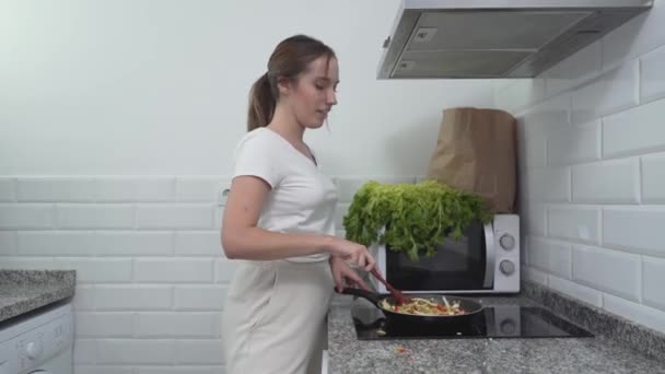 Mujer joven cocina plato de verduras en la estufa de gas eléctrico en su apartamento. - tiro medio — Vídeos de Stock