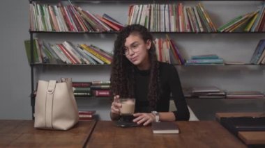 Young Girl Drinks Coffee Then Read A Book On Wooden Table WIth Book Shelf In Background. - static shot