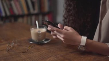 Hands Of Young Woman Typing On Smartphone With A Cup Of Creamy Coffee And Eyeglasses Lying On The Table. close up