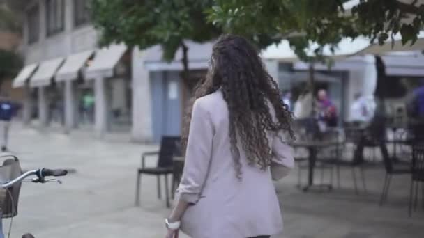 Chica marroquí con abrigo y pelo largo rizado caminando en la calle junto con un hombre empujando una bicicleta. - tiro de seguimiento — Vídeo de stock