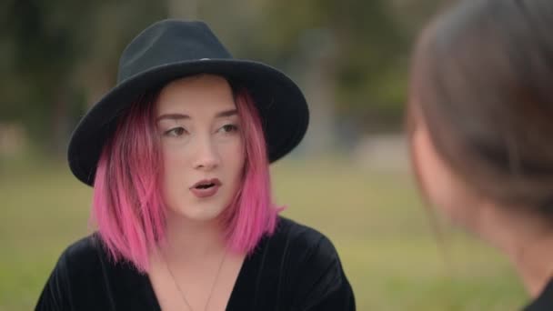 Bonita chica de pelo rosa usando un sombrero negro habla con otra chica. de cerca, sobre el hombro disparado — Vídeos de Stock