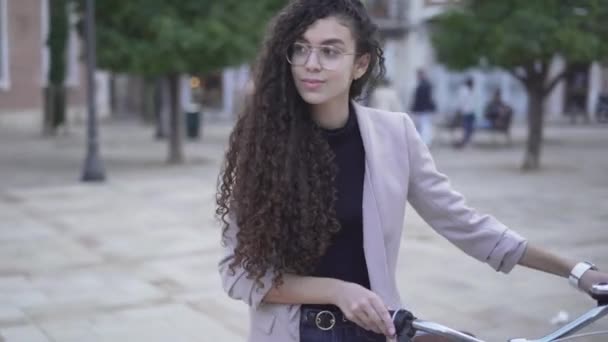 Charmig ung kvinnlig modell i glasögon med långa lockigt hår promenader med en cykel utomhus. medelstora skott — Stockvideo