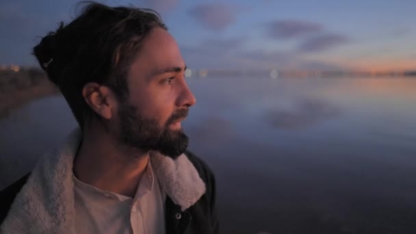 Porträtt av en stilig skäggig man som njuter av en solnedgång vid sjön och tittar på kameralinsen, blinkar med ögonen och slät skägg - slow motion — Stockvideo