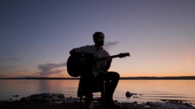 Gölün kenarındaki sandalyede oturan gitar çalan bir adamın silueti. Renkli bir günbatımı arka planında. 