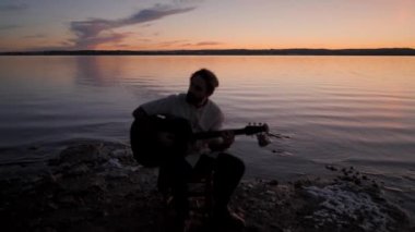 İspanya 'da dramatik günbatımında göl kenarında akustik gitar akorları çalan bir gitaristin silueti. 