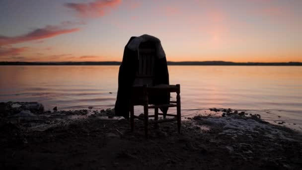 Silhuett av en trästol med hängande jacka vid sjön på färgglad solnedgång - gimbal pullback — Stockvideo