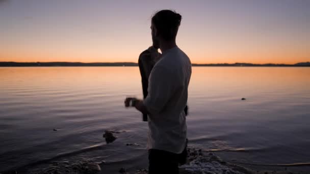 Silhuett av skäggig hipster man sätta på sin höst jacka står vid sjön vid solnedgången — Stockvideo